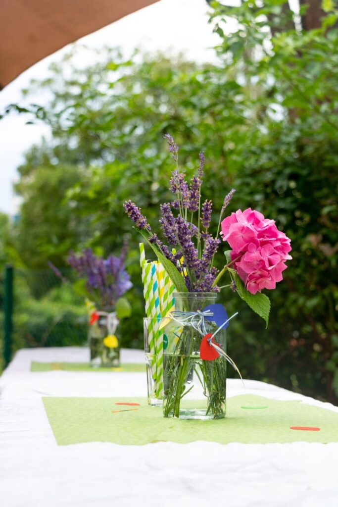 Zarter Blumenschmuck in einem Glasgefäß auf einem Tisch, umgeben von Bäumen im weichen Tageslicht.