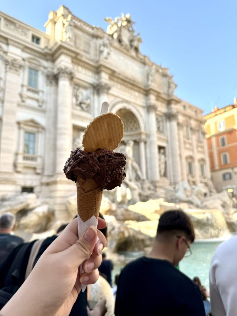 Eiswaffel im Vordergrund vor dem Trevi-Brunnen in Rom, im Hintergrund Besucher und barocke Architektur.
