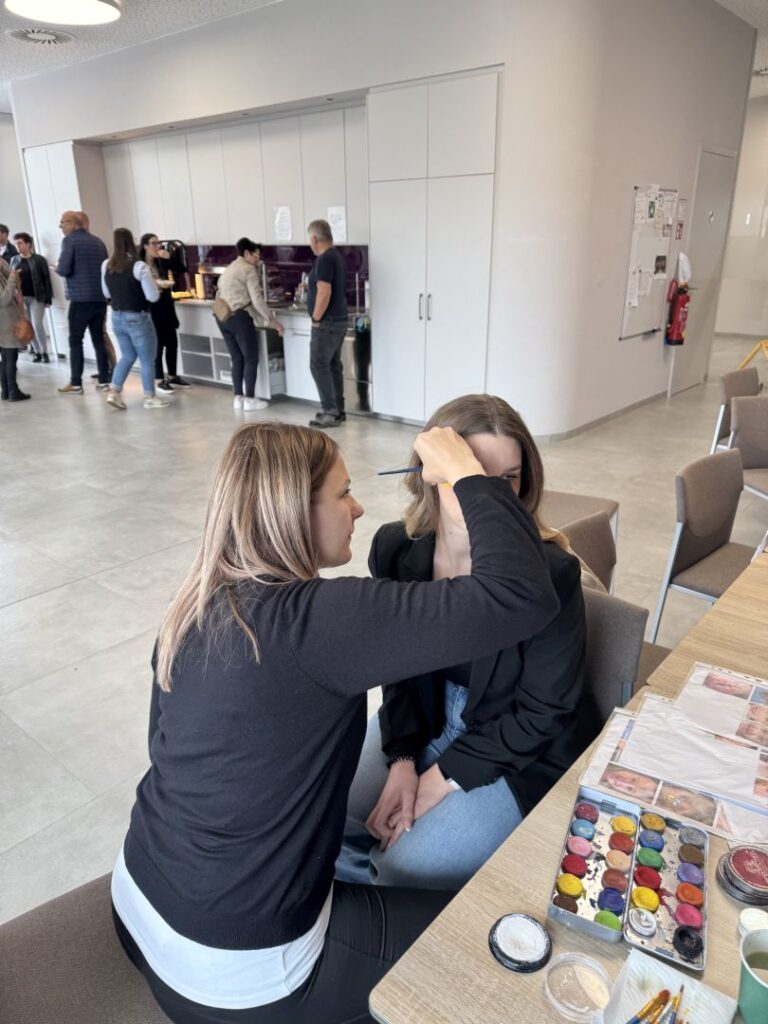 Kinderschminken: Eine Person schminkt eine sitzende Frau, Farbenpalette am Tisch
