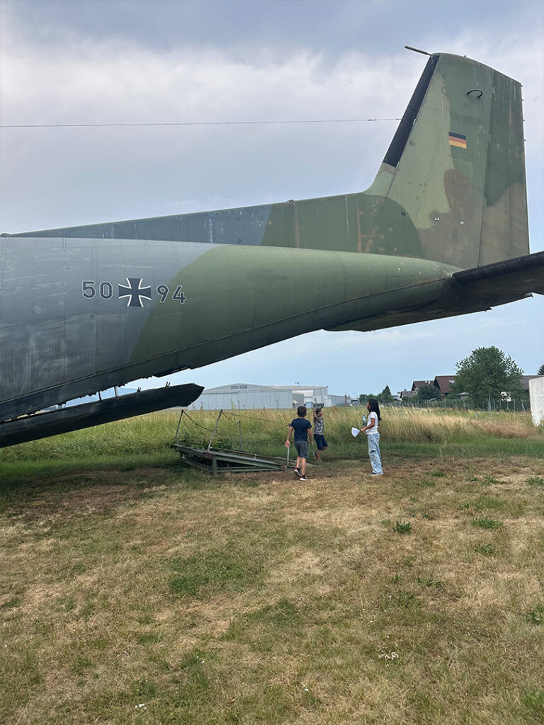 Rückansicht eines großen grünen Militärtransportflugzeugs mit Tarnlackierung, davor stehen drei Personen auf einem grasigen Feld neben dem Flugzeug.