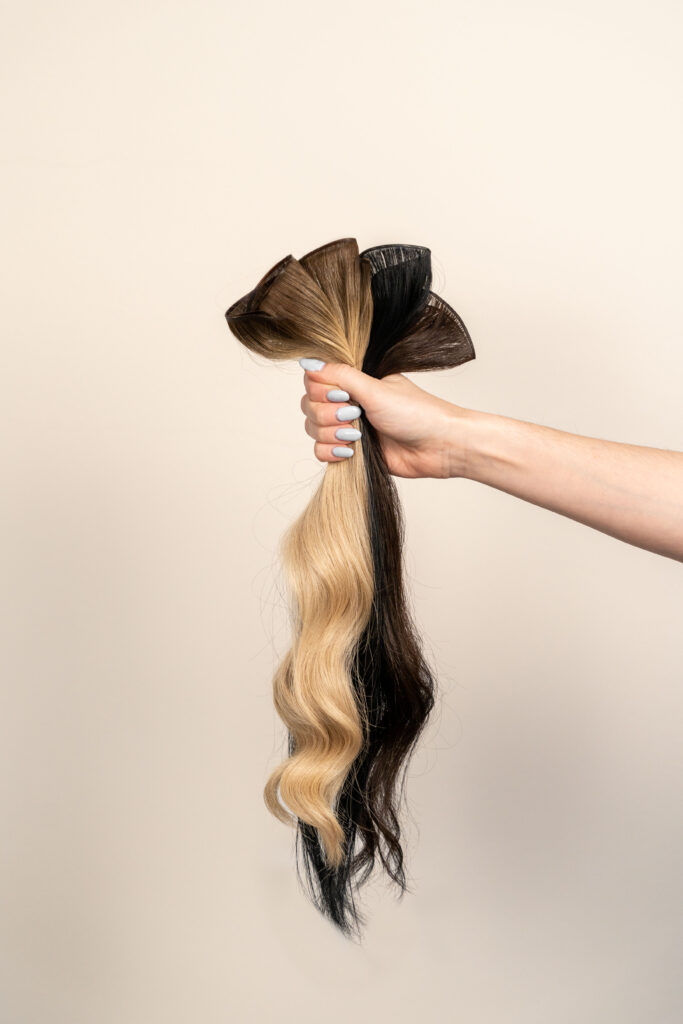 Hand hält verschiedene Echthaar-Extensions in Blond-, Braun- und Schwarztönen vor hellem Hintergrund.